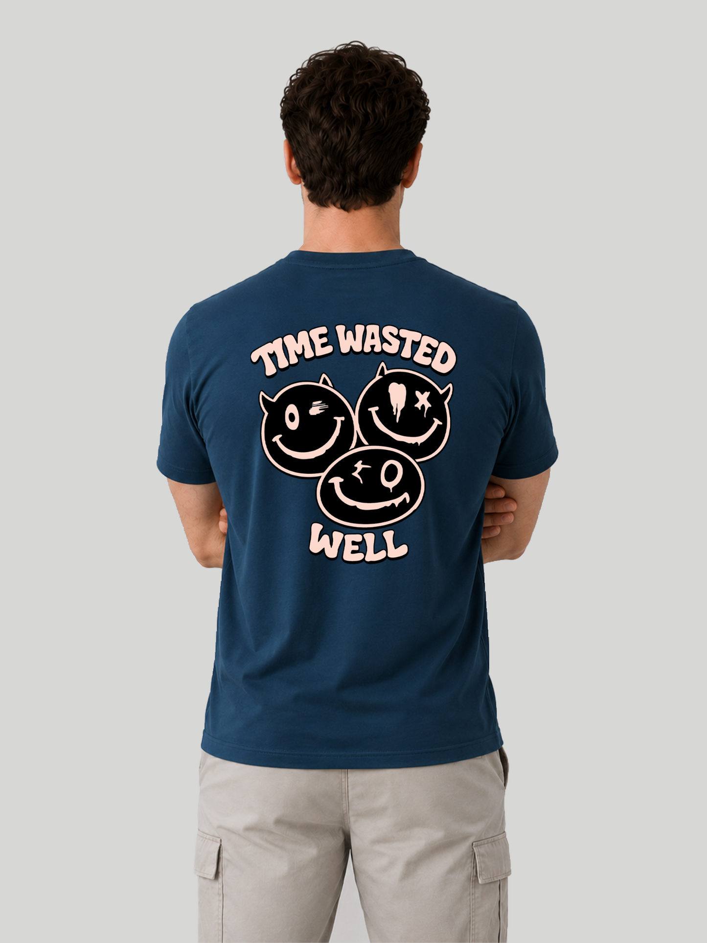 The back view of a male model standing with arms crossed, wearing a navy blue regular fit t-shirt with a light pink "TIME WASTED WELL" typography and edgy smiley print across the back.