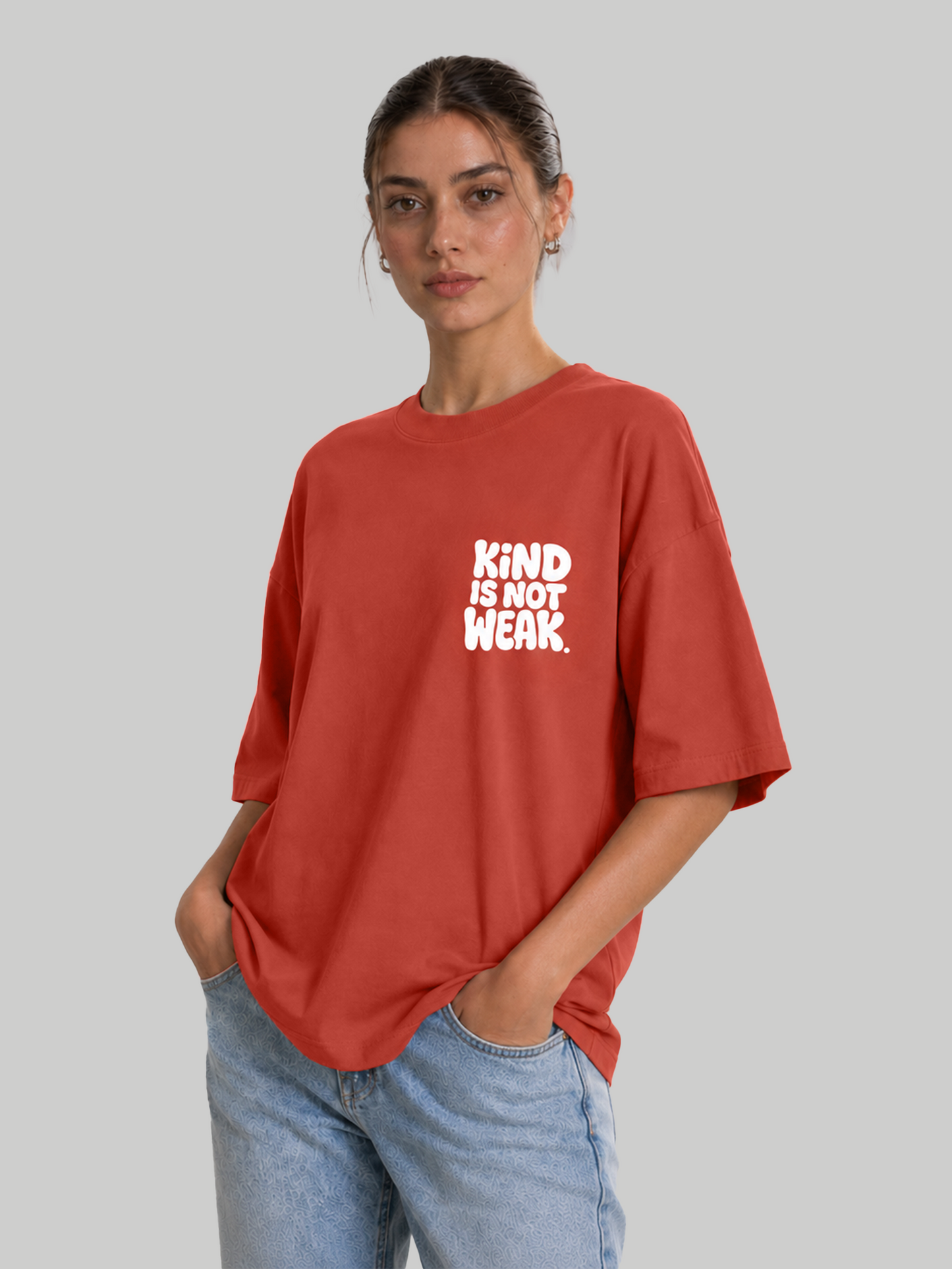 Women's oversized brick red graphic t-shirt with "Kind Is Not Weak" design, model standing with hands in pockets.