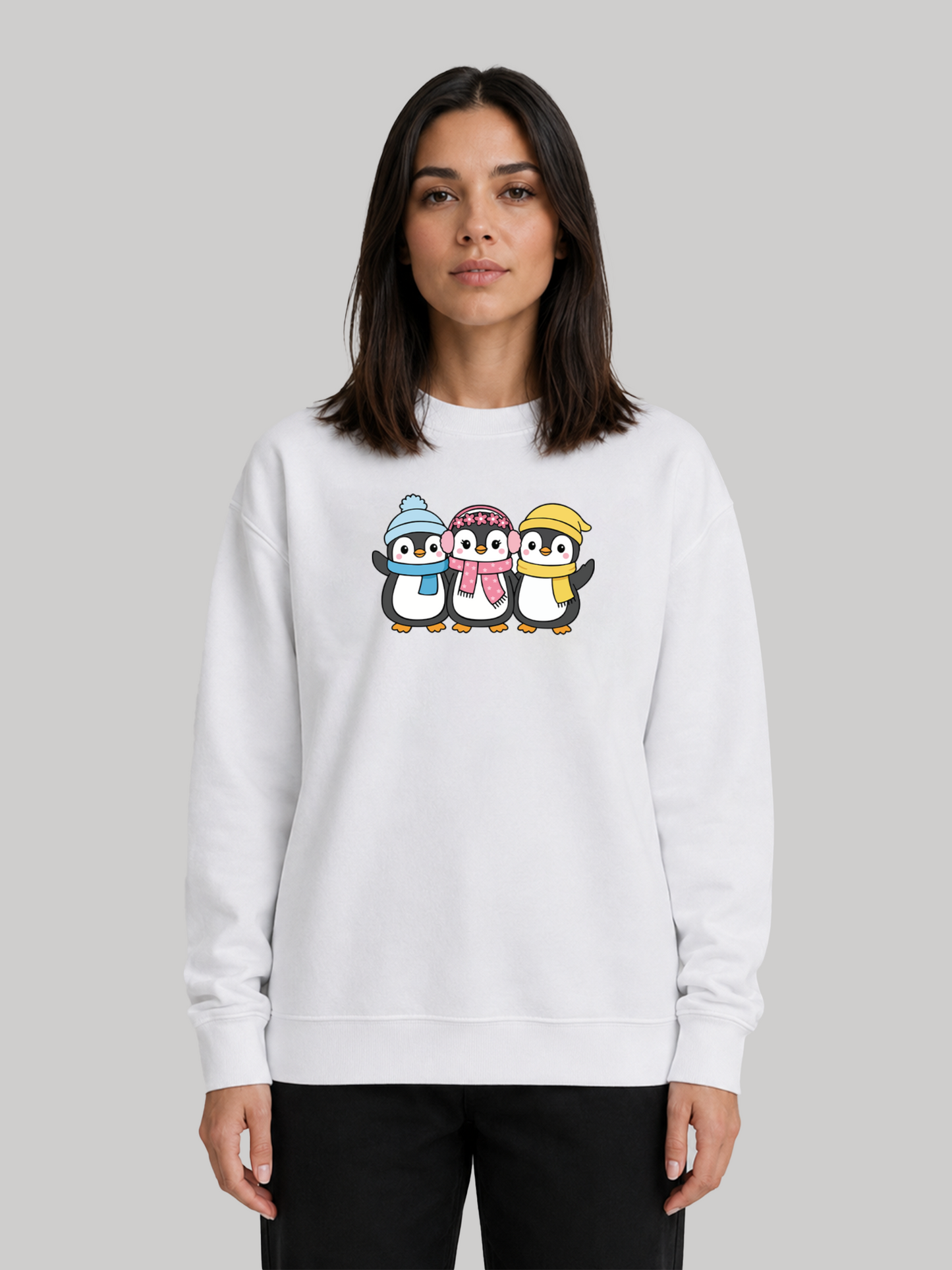 A straight front view of a female model wearing a white crewneck sweatshirt with a cute graphic print of three cartoon penguins wearing winter hats and scarves.