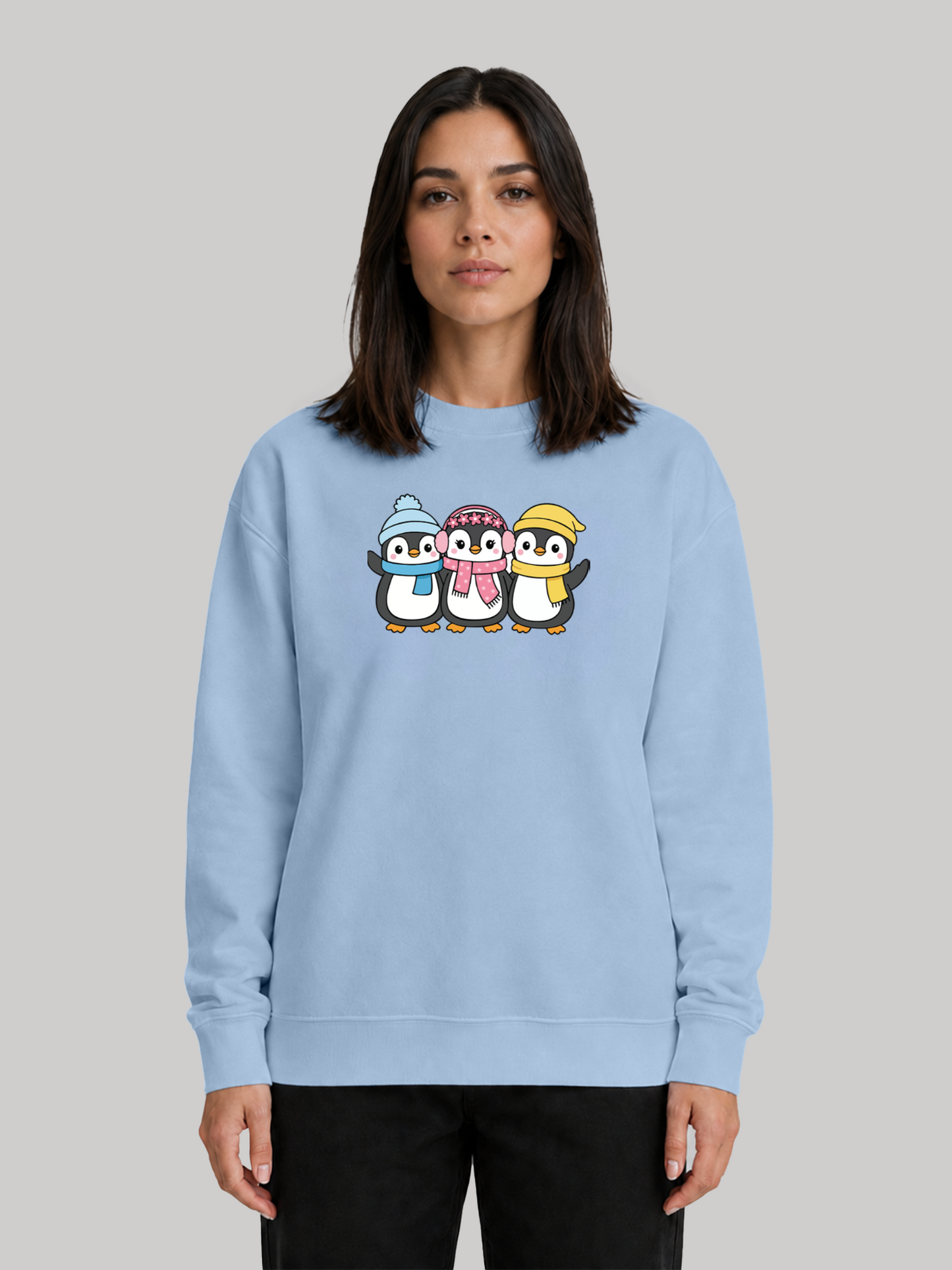 A full front view of a female model wearing a baby blue sweatshirt with a colorful graphic print of three cute winter penguins across the chest.