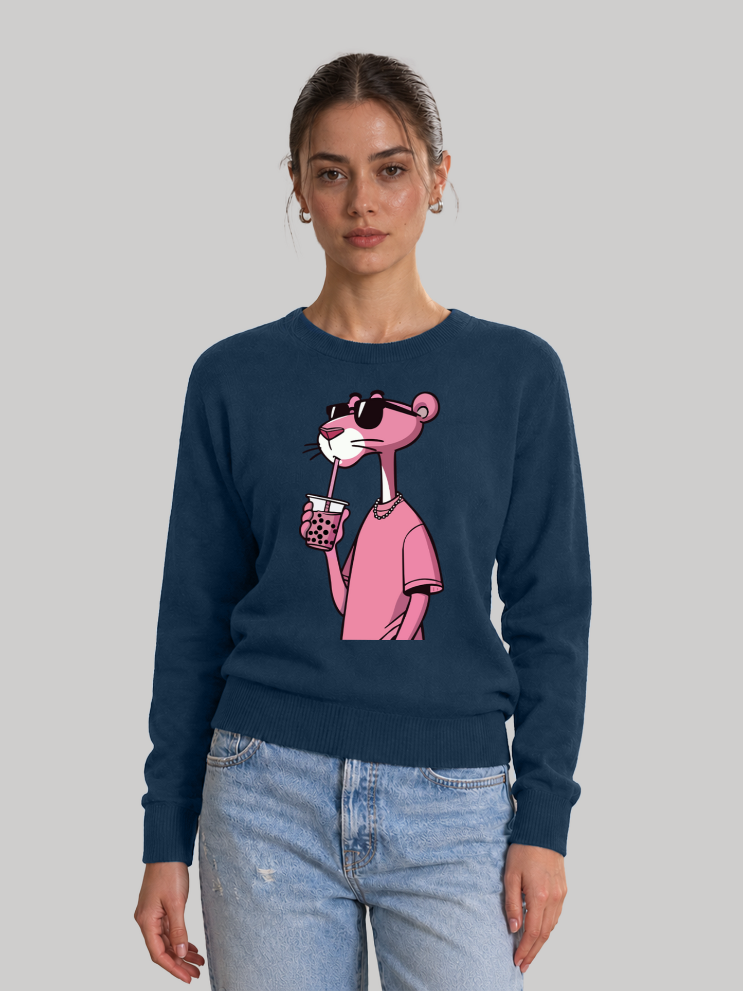 A straight front view of a female model wearing a navy blue crewneck sweatshirt with a pink cartoon panther graphic drinking boba tea, paired with light blue jeans.