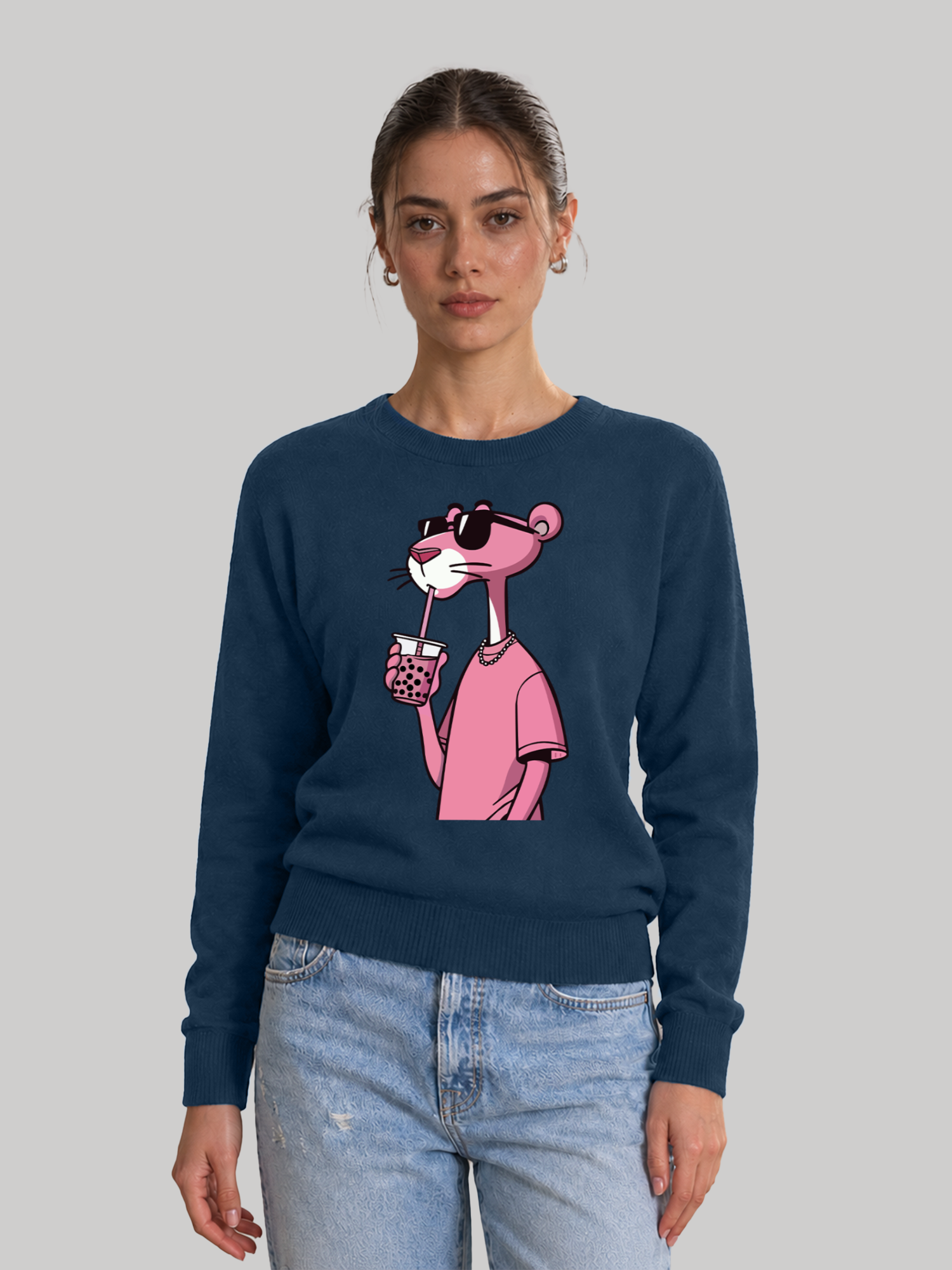 A straight front view of a female model wearing a navy blue crewneck sweatshirt with a pink cartoon panther graphic drinking boba tea, paired with light blue jeans.