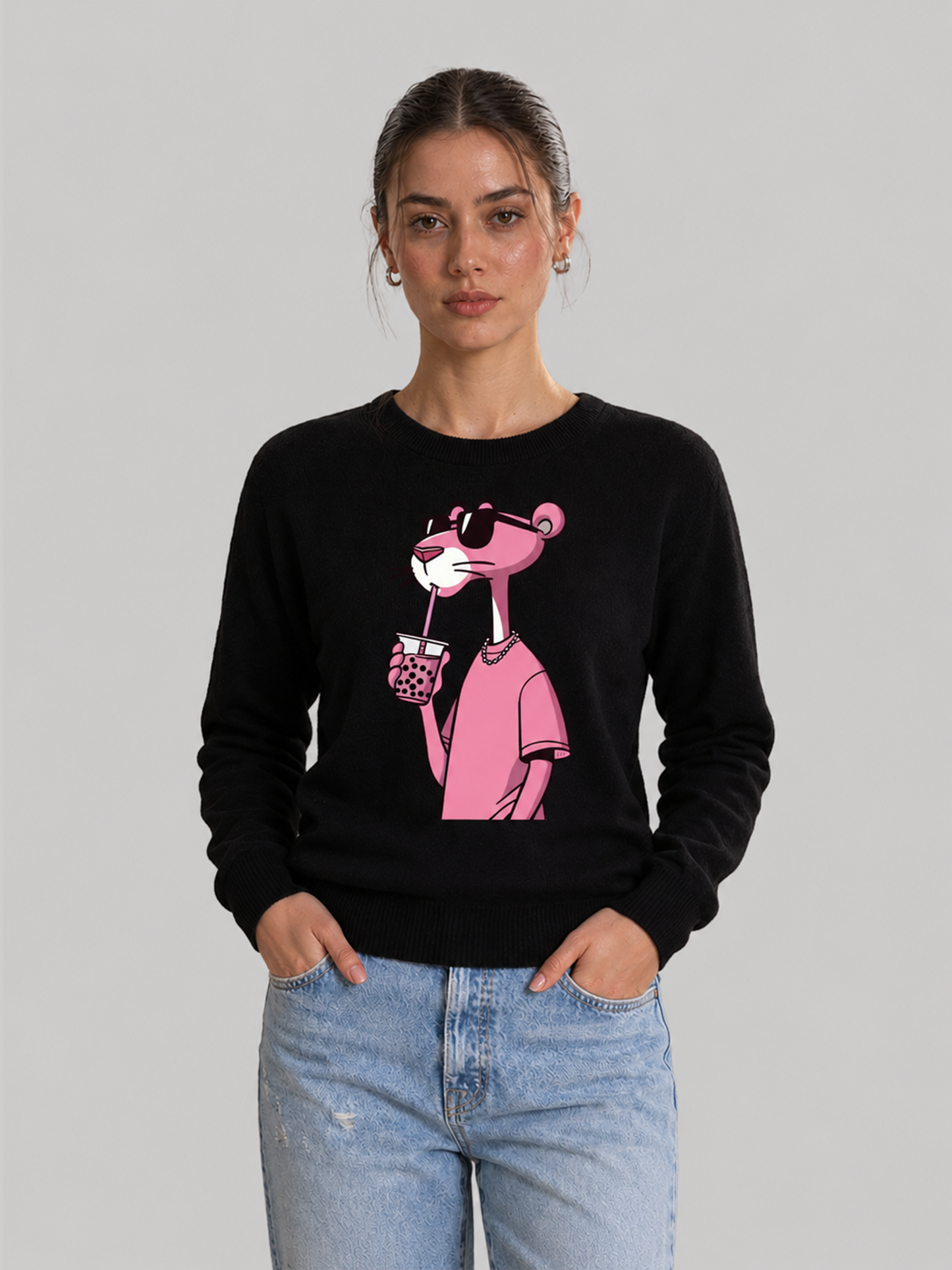A front view of a female model standing with her hands in her pockets, wearing a black crewneck sweatshirt with a graphic print of the Pink Panther wearing sunglasses and drinking boba tea.