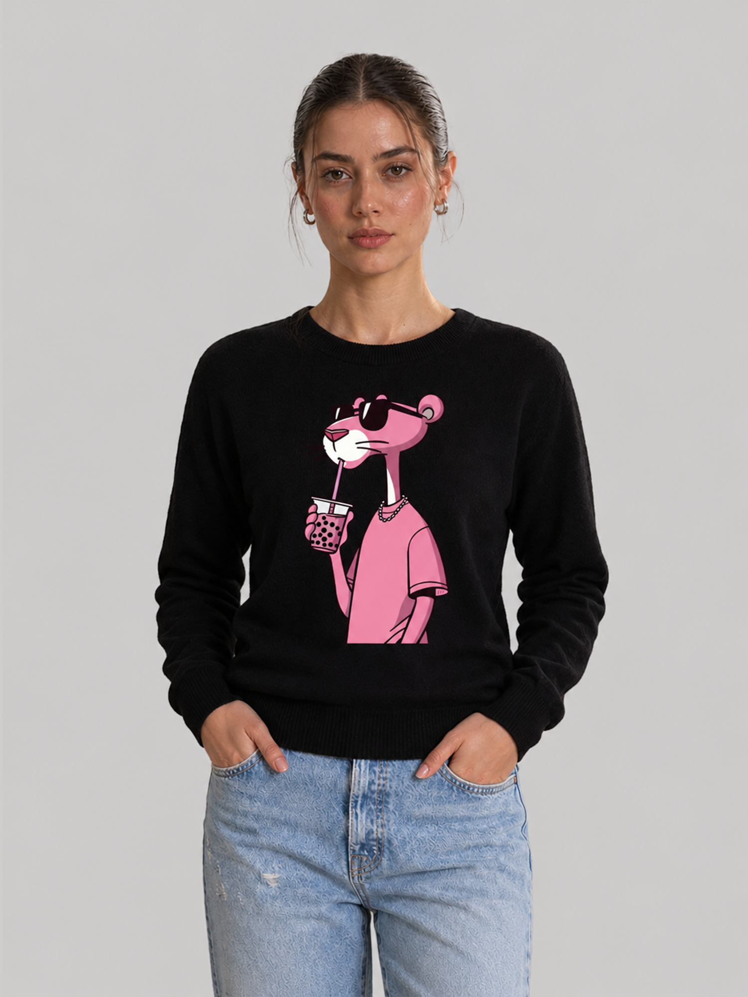 A front view of a female model standing with her hands in her pockets, wearing a black crewneck sweatshirt with a graphic print of the Pink Panther wearing sunglasses and drinking boba tea.
