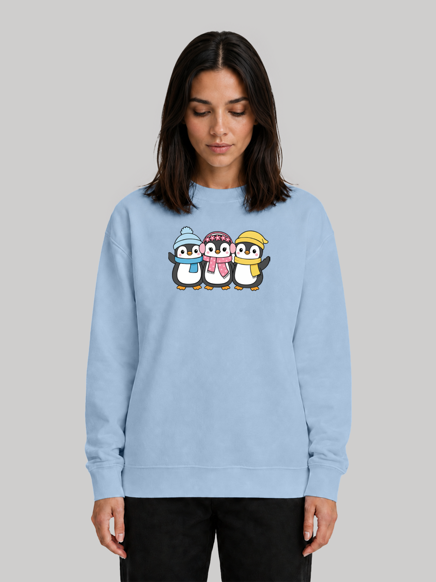 A female model looking down slightly, showcasing a baby blue long-sleeve crewneck sweatshirt with a playful cartoon penguin graphic.