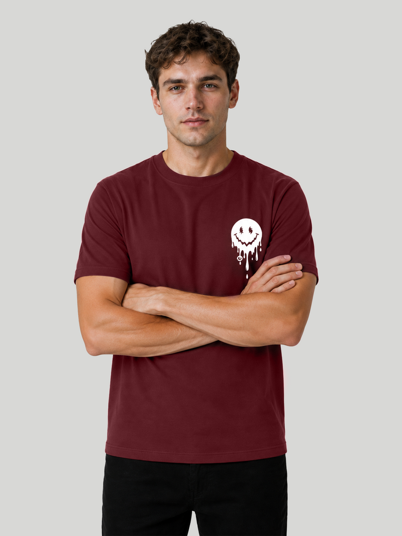 A front view of a male model standing with his arms crossed, wearing a maroon crewneck half-sleeve t-shirt with a small white melting smiley face graphic printed on the left chest.