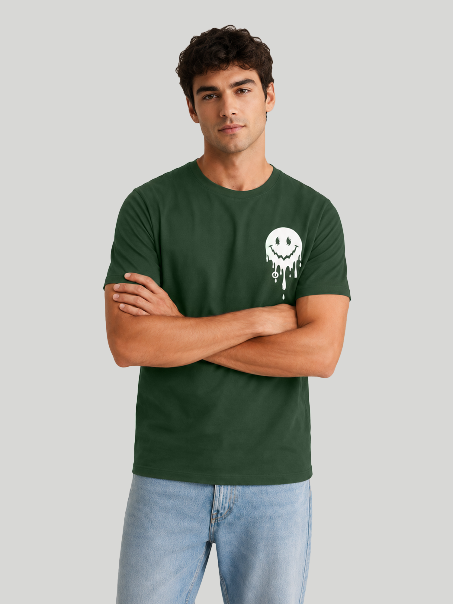 A front view of a male model standing with arms crossed, wearing an olive green regular fit t-shirt with a small white dripping smiley graphic on the chest, paired with light blue jeans.