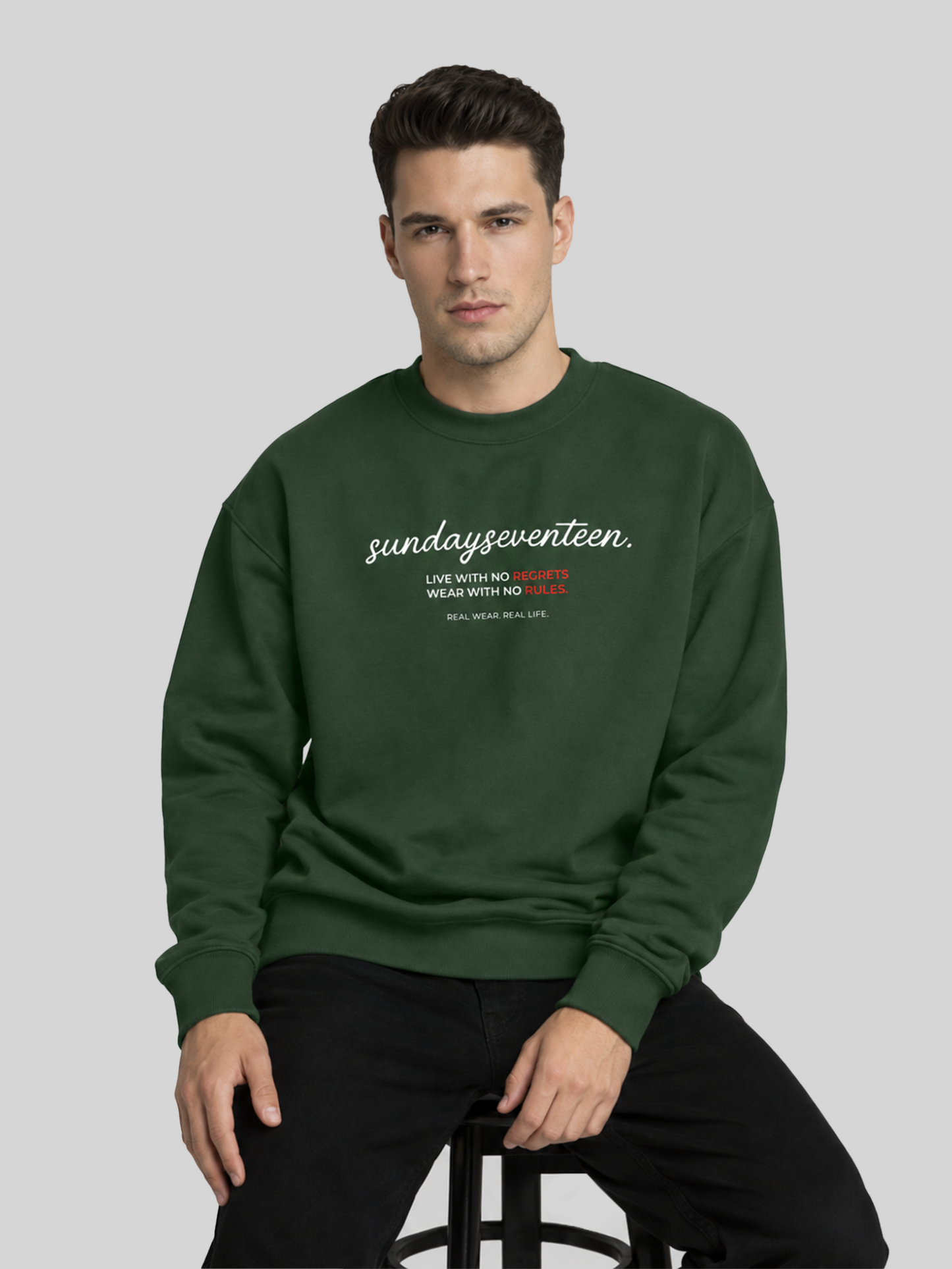 A relaxed pose of a male model sitting and wearing an olive green graphic sweatshirt paired with black trousers.