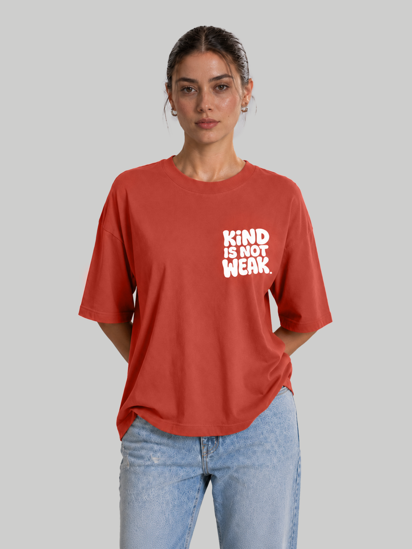 Women's oversized brick red t-shirt with "Kind Is Not Weak" white chest print, hands behind back.