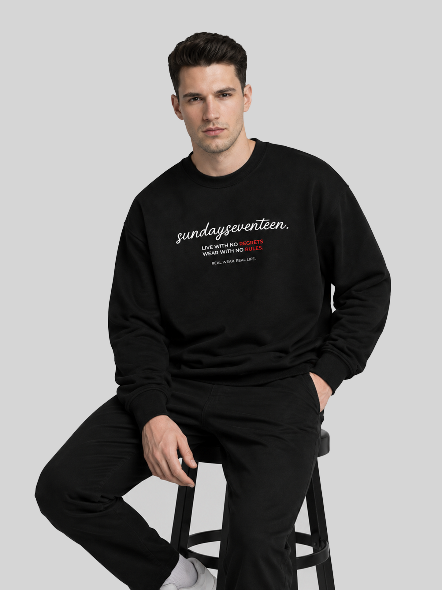 A male model sitting on a stool wearing a black long-sleeve sweatshirt with white text "sundayseventeen" and black pants, showcasing a casual streetwear look.