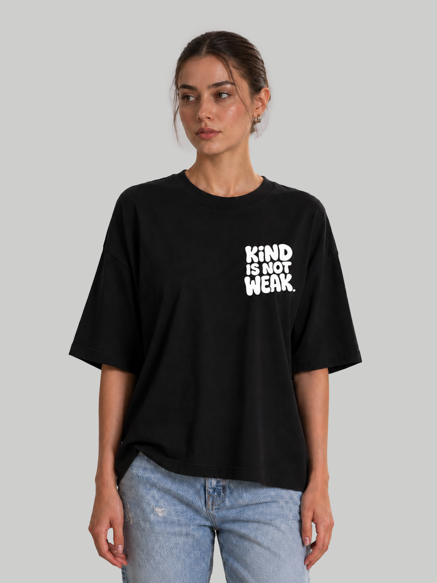 Women's oversized black t-shirt with "Kind Is Not Weak" white chest print, front-facing model looking sideways.