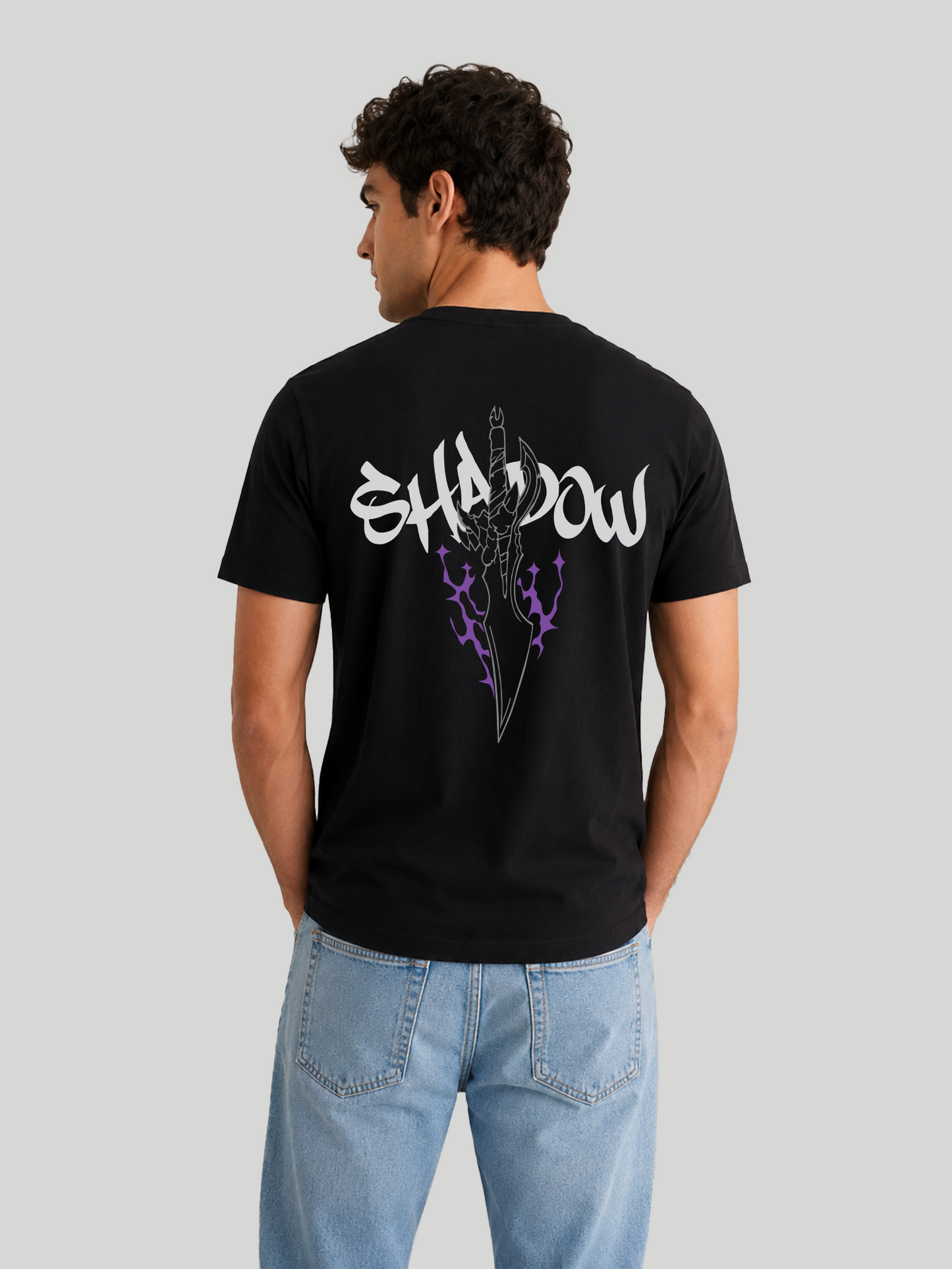 The back view of a male model looking slightly over his shoulder, wearing a black cotton half-sleeve t-shirt with a bold white typography print reading "SHADOW" and an edgy purple flame dagger graphic across the back.