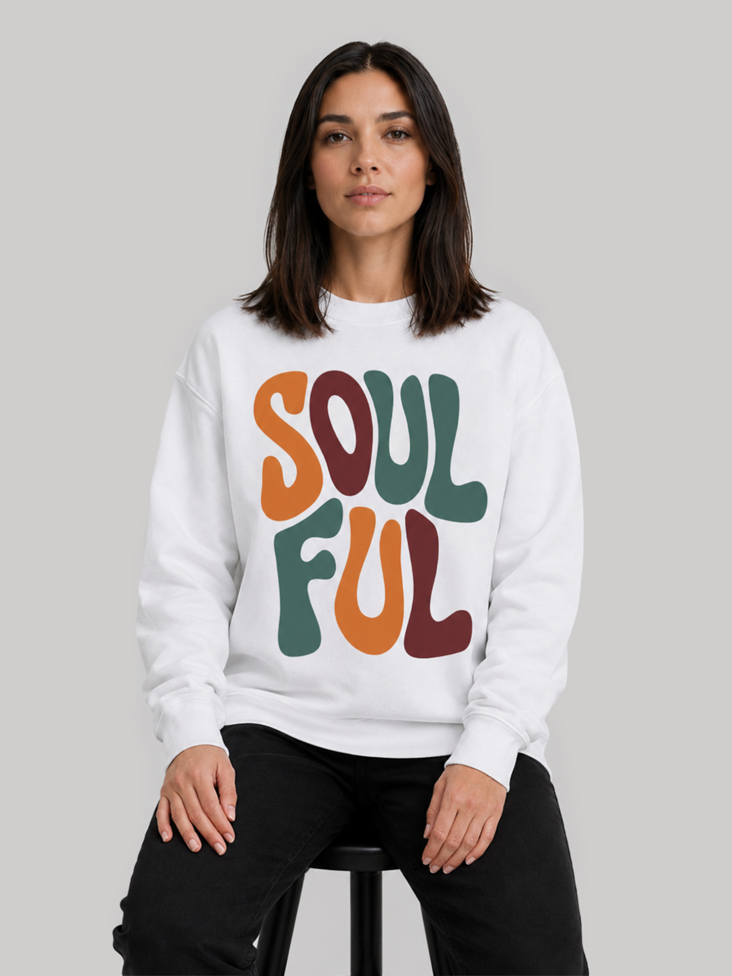A female model sitting on a stool wearing a white streetwear sweatshirt with a bold typography graphic print and black trousers.