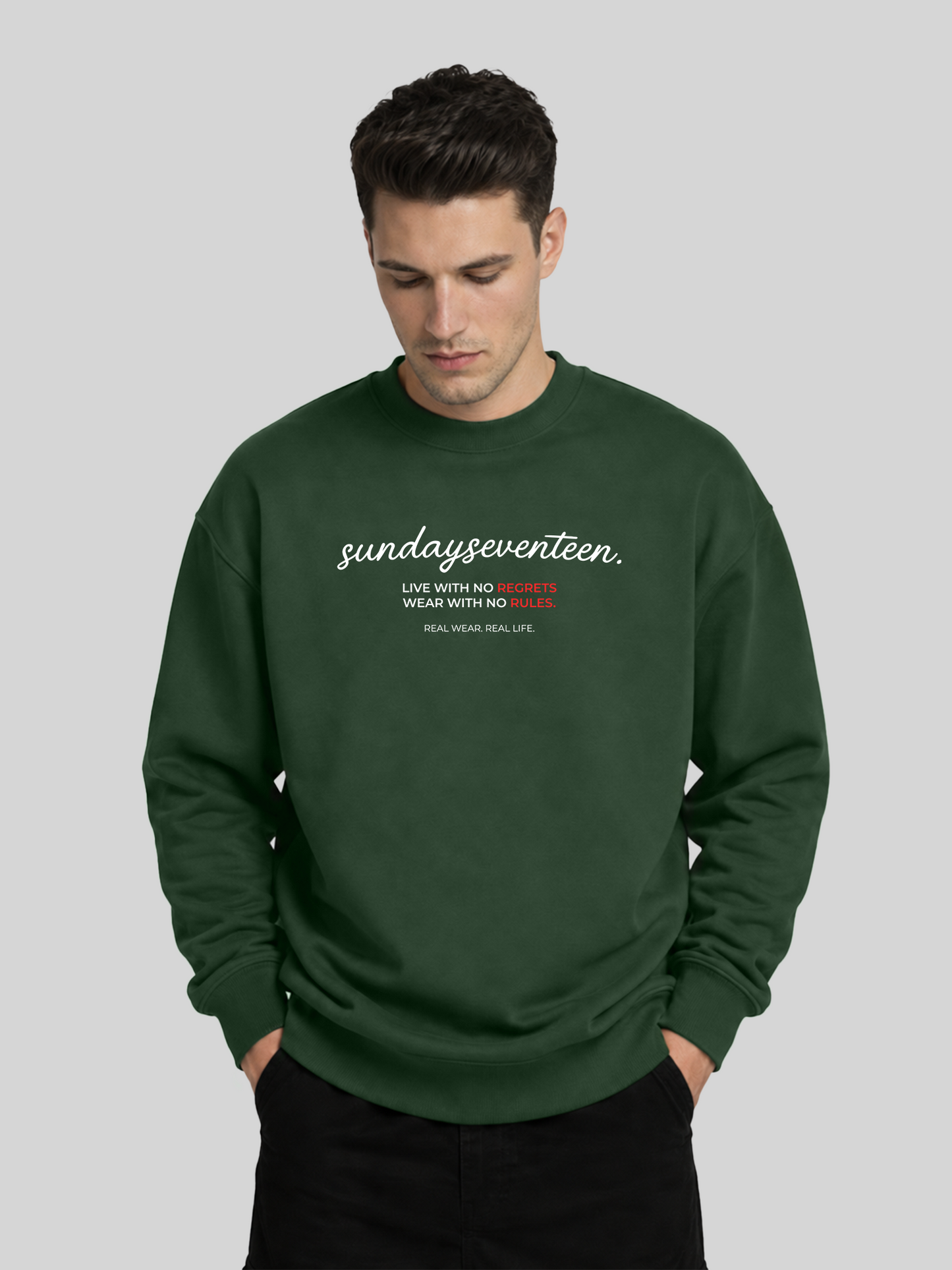 A male model standing in an olive green long-sleeve sweatshirt looking slightly down, showcasing the comfort and premium fabric texture.