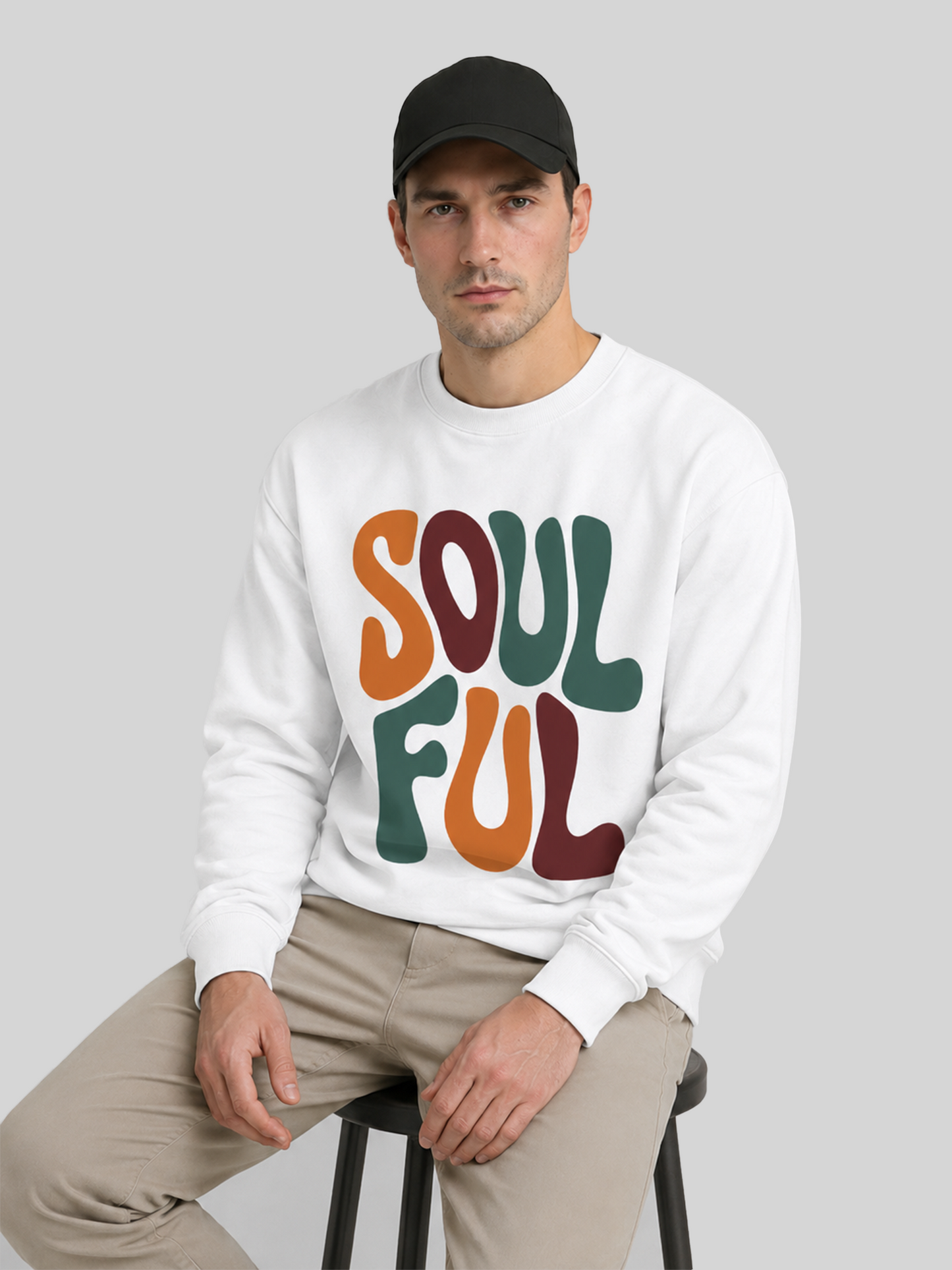 A front view of a male model standing with his arms crossed, wearing a black baseball cap and a white long-sleeve sweatshirt with a retro typography design.