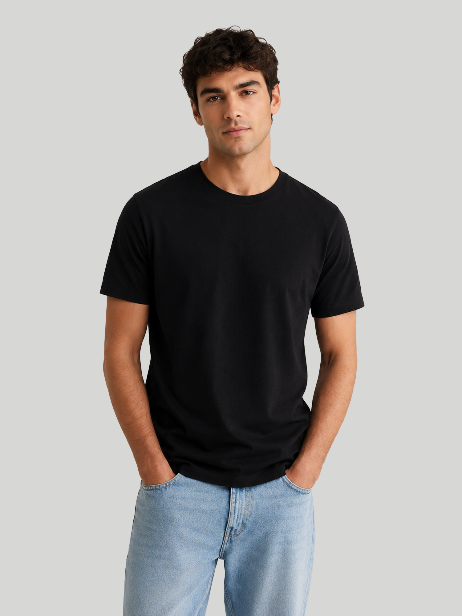 A front view of a male model looking forward with both hands in his pockets, wearing a plain black premium cotton regular fit round neck t-shirt paired with light blue jeans.