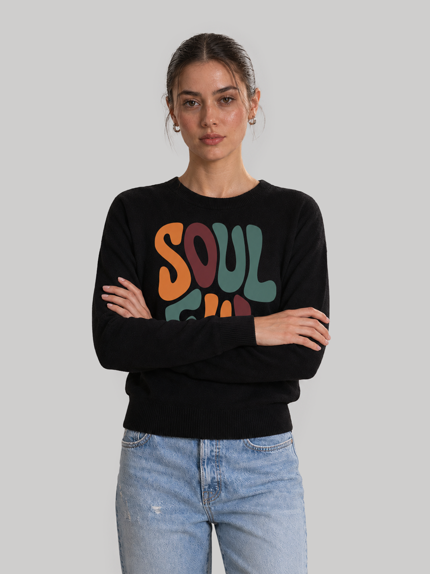 A front view of a female model standing with arms crossed, wearing a black crewneck sweatshirt featuring a colorful retro "SOULFUL" typography print paired with light wash denim jeans.