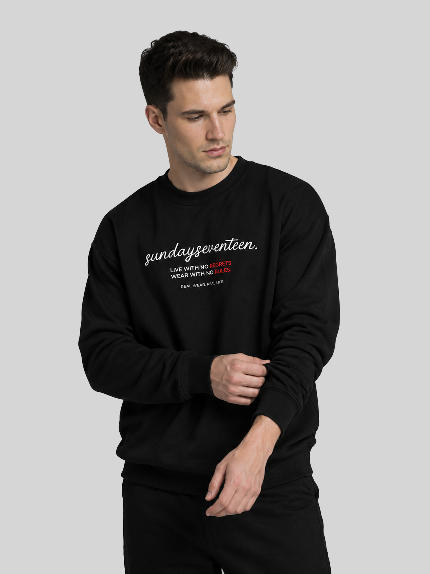 A male model looking down while adjusting the sleeve of a black crewneck sweatshirt featuring a graphic typography print on the chest.