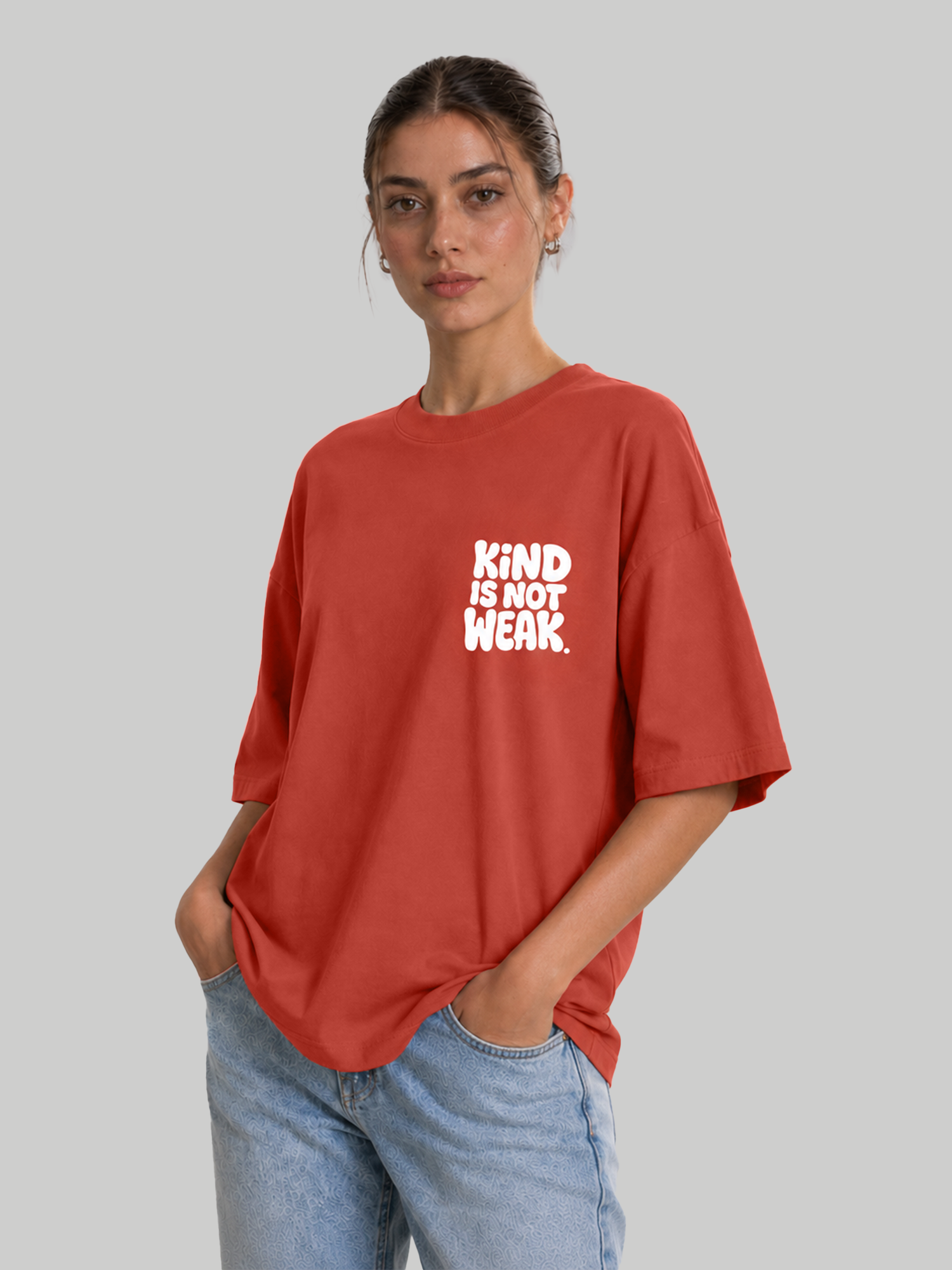 Women's oversized brick red graphic t-shirt with "Kind Is Not Weak" design, model standing with hands in pockets.